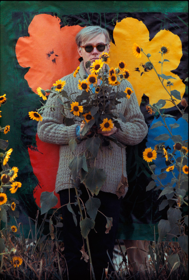 Andy-Warhol-in-a-field-of-black-eyed-Susans-holding-a-bouquet-of-flowers-with-an-early-_Flowers_-canvas-serving-as-a-backdrop-in-Queens,-New-York
